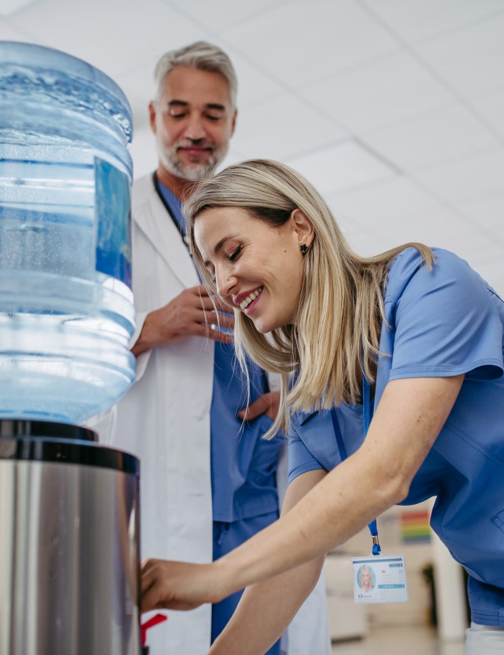 Doctor and nurse taking a break during work shift at hospital, drinking water from water dispenser in hospital lobby, talking. Handsome doctor and nurse taking a break during work shift at hospital, drinking water from water dispenser in hospital lobby, talking.